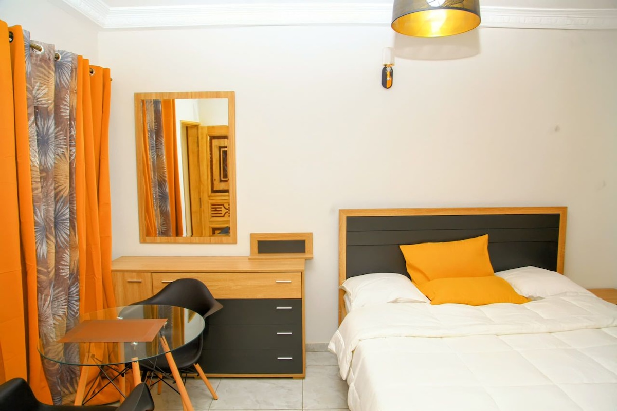 A neatly arranged room features a spacious bed with a vibrant orange accent pillow. A round glass table and black chairs are positioned nearby, while a wooden dresser reflects natural light from a nearby window. Orange curtains add a pop of color to the neutral walls.