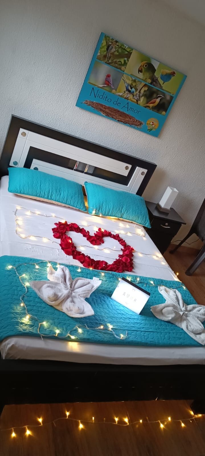 A spacious bed is adorned with a turquoise quilt and decorated with rose petals arranged in a heart shape. Soft white towels are shaped like swans at the foot of the bed. Fairy lights add a gentle glow to the inviting bedding.