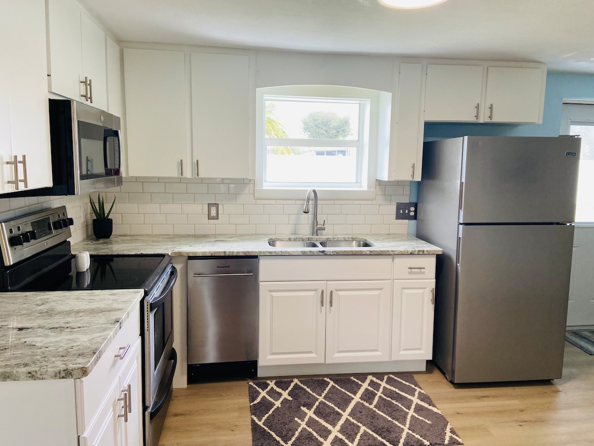 A bright kitchen area features modern appliances, including a stainless steel refrigerator and an electric stove. The white cabinetry and light countertops are complemented by a patterned rug on the floor. Natural light enters through a window, enhancing the space.
