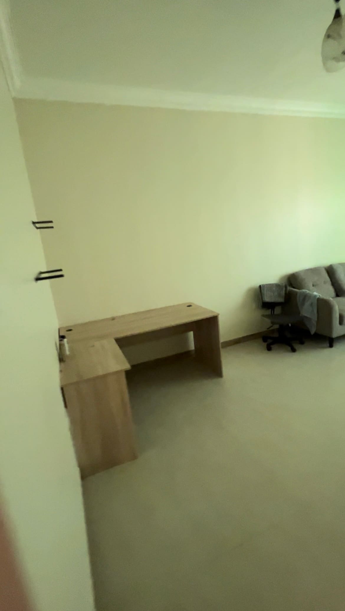 A bright and spacious room contains a simple wooden desk paired with a comfortable chair. A grey sofa is positioned against the wall, providing an inviting space for relaxation. The walls are painted in a soft, neutral tone, emphasizing the room's openness.