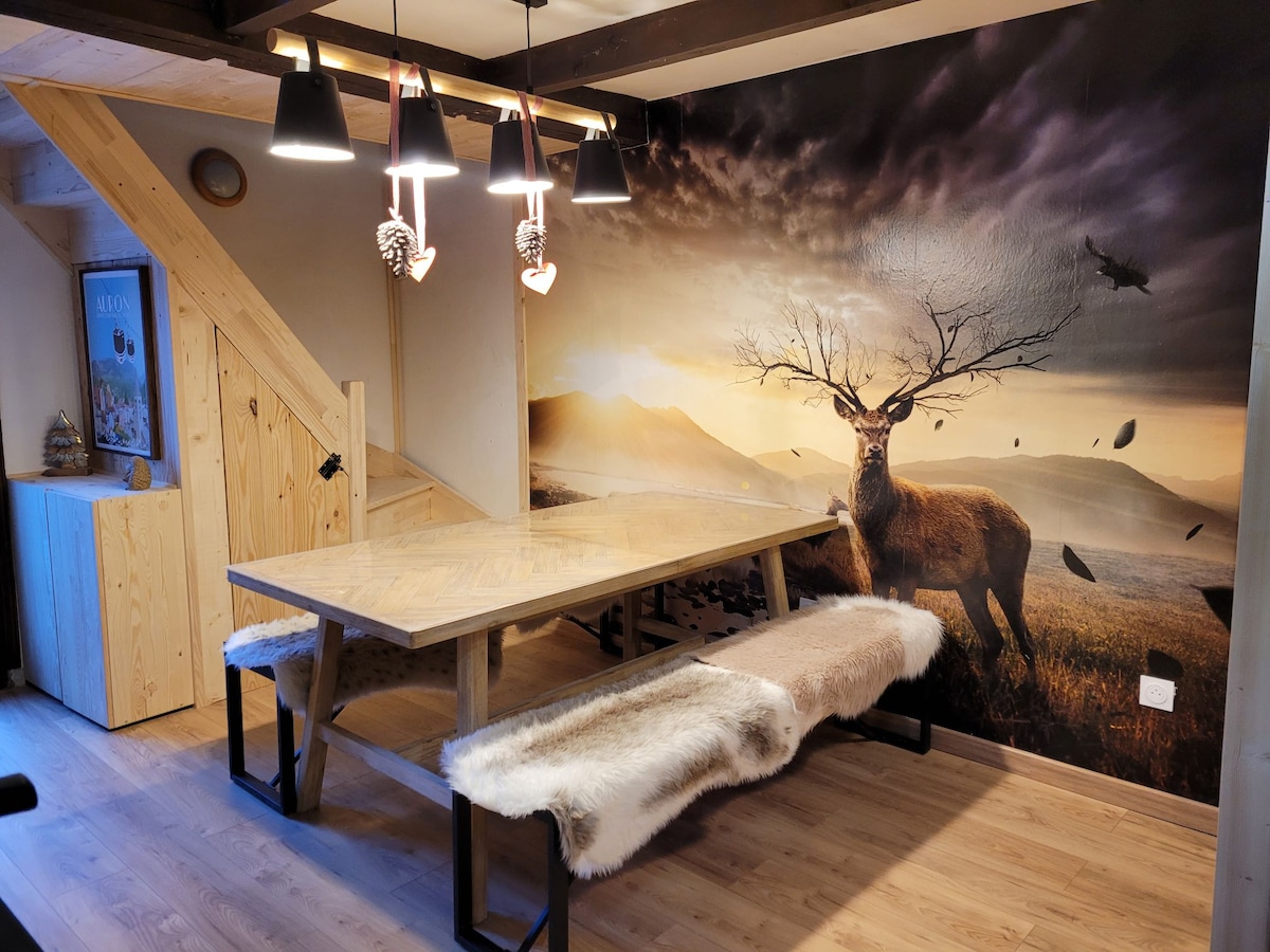 A dining area is presented with a wooden table and benches, featuring soft fur accents. The wall behind displays a large mural of a landscape with a deer, complemented by pendant lights above, creating a cozy gathering space.