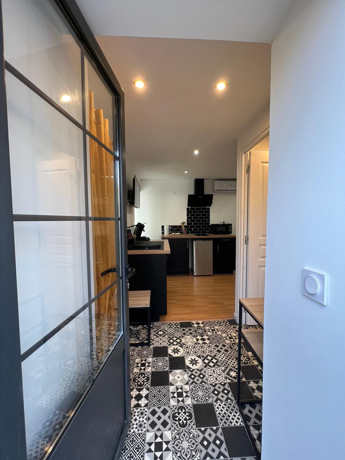 A modern entryway is presented, featuring a patterned tile floor in black and white. A glass-paneled door allows for light to enter, with glimpses of a kitchen area visible in the background. Contemporary appliances and cabinetry are discernible in the space.