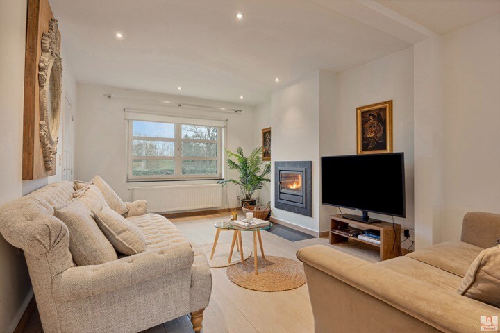 A cozy living area features a light-colored sofa and a plush armchair, arranged around a round coffee table. A modern fireplace is positioned beneath a wall-mounted TV. Soft lighting enhances the neutral tones throughout the space, while a large window offers views of the surrounding landscape.