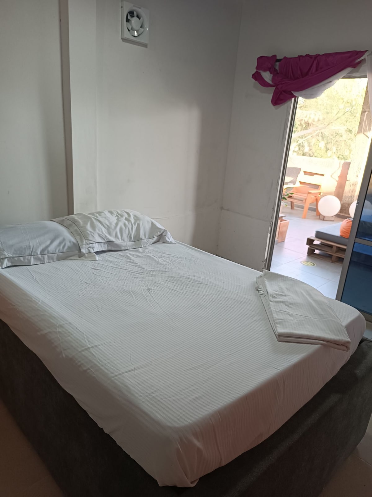 A cozy sleeping area is shown, featuring a spacious bed dressed in crisp white bedding. Natural light enters through an open door, leading to a small outdoor space furnished with seating. A neatly folded towel rests on the bed's edge.