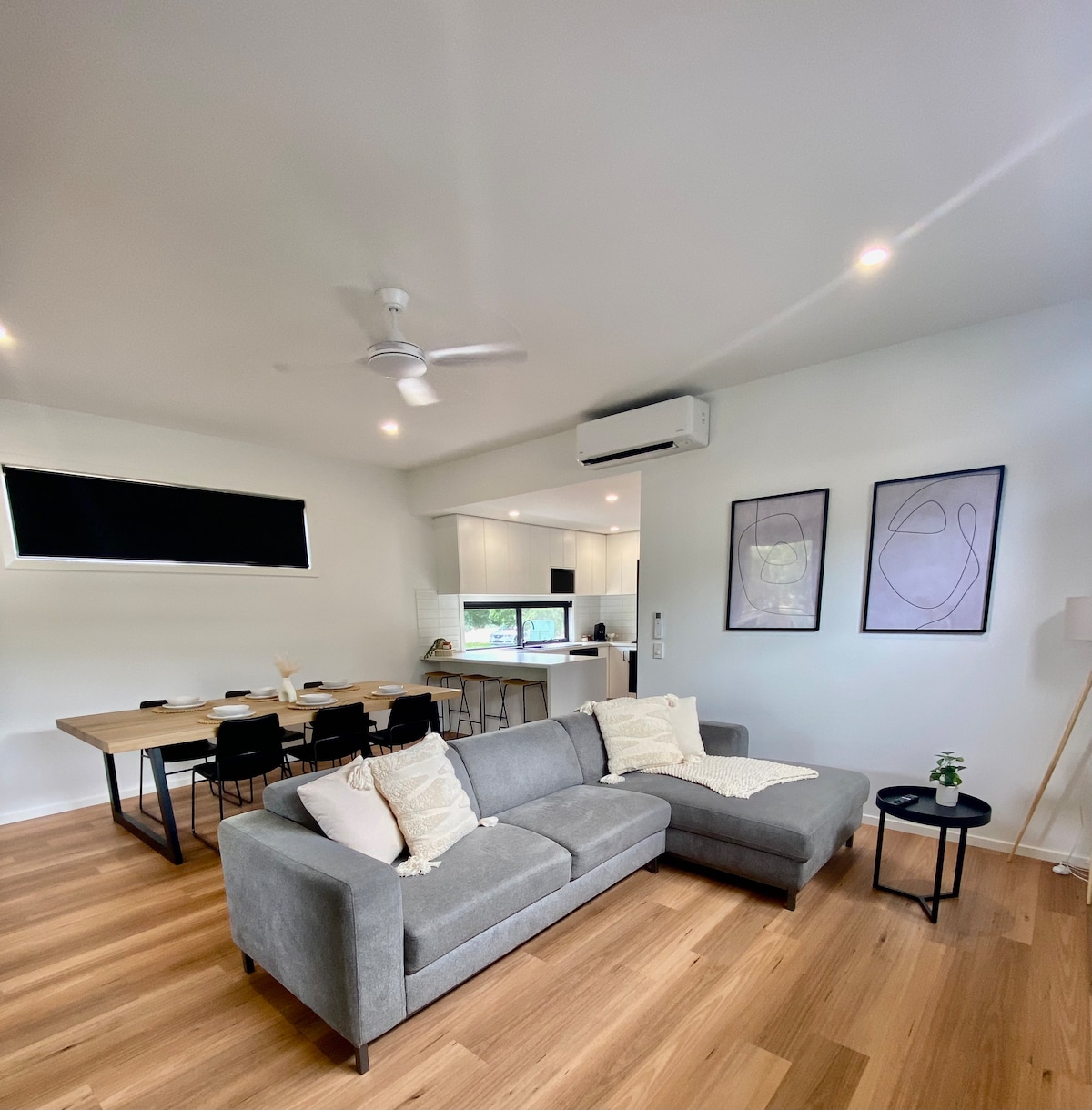 An open-plan living area features a modern sectional sofa with decorative cushions. A dining table is visible in the background, accompanied by black chairs. Natural light filters through large windows, and a minimalist ceiling fan is positioned above the seating area.