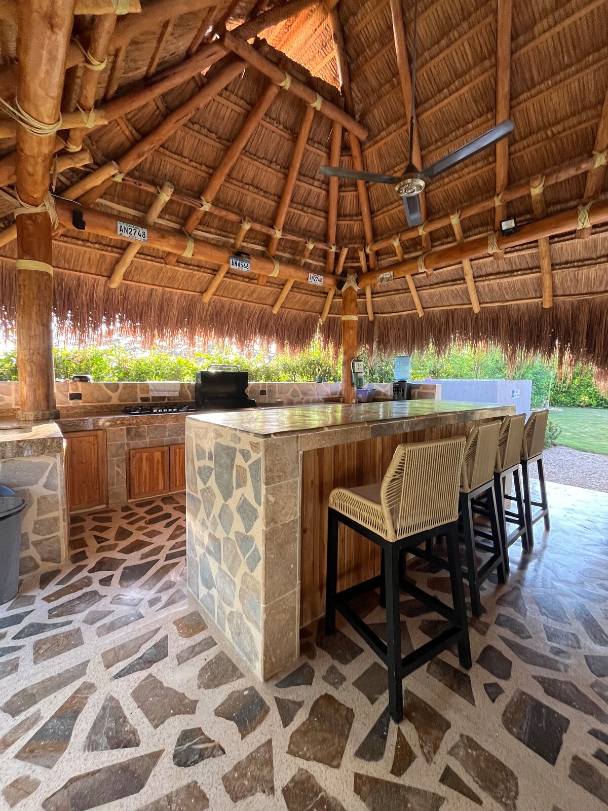 An outdoor kitchen space features a thatched roof supported by wooden beams. A stone counter with wooden cabinetry holds a cooking area and a sink. Four high-backed stools are positioned around the counter, with natural stone flooring enhancing the rustic design.