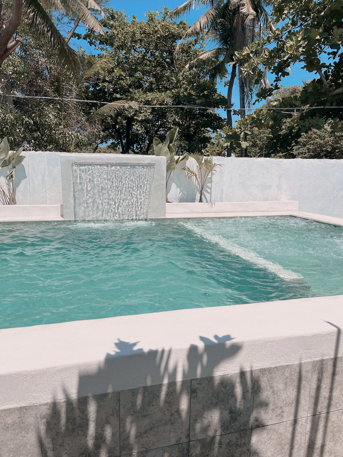 A refreshing swimming pool is depicted with a waterfall feature cascading into the clear water. Surrounding palm trees provide shade, while the stucco walls create a serene atmosphere. Sunlight glimmers on the water's surface, enhancing the tranquil setting.