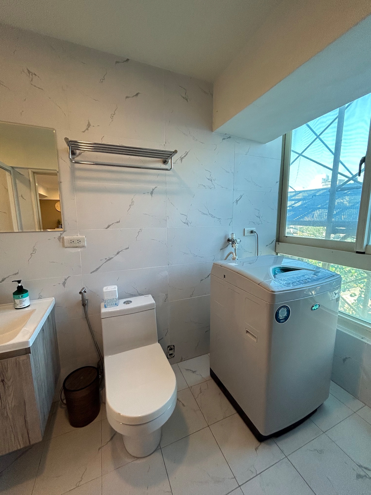 The bathroom features a compact design with functional elements. A toilet is positioned next to a washing machine, while a mirror above a sink adds practicality. Natural light enters through a window, illuminating the room's minimalist decor.