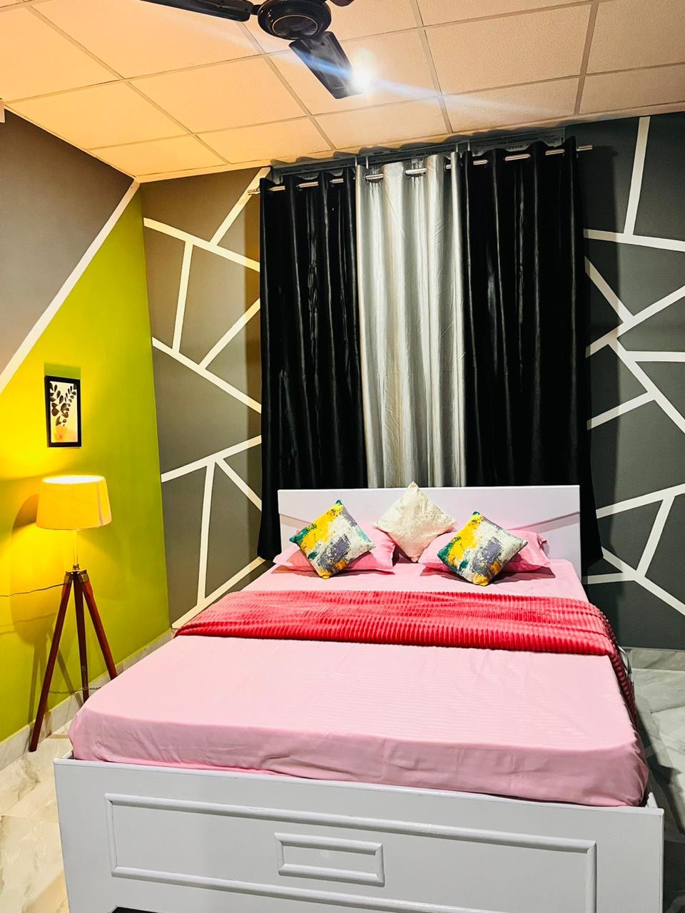 A comfortable sleeping area is presented with a spacious bed dressed in light pink bedding. Brightly colored pillows add a pop of color against the geometric patterned walls. A standing lamp provides additional lighting, while dark curtains frame the window.