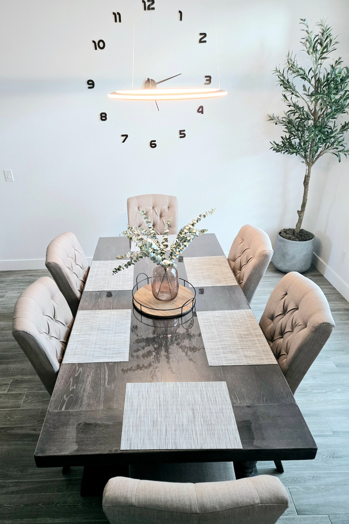 A spacious dining table is surrounded by six upholstered chairs, offering a comfortable setting for meals. A decorative centerpiece with greenery is placed on the table. A minimalist wall clock adds a modern touch, while a potted plant in the corner provides a hint of nature.