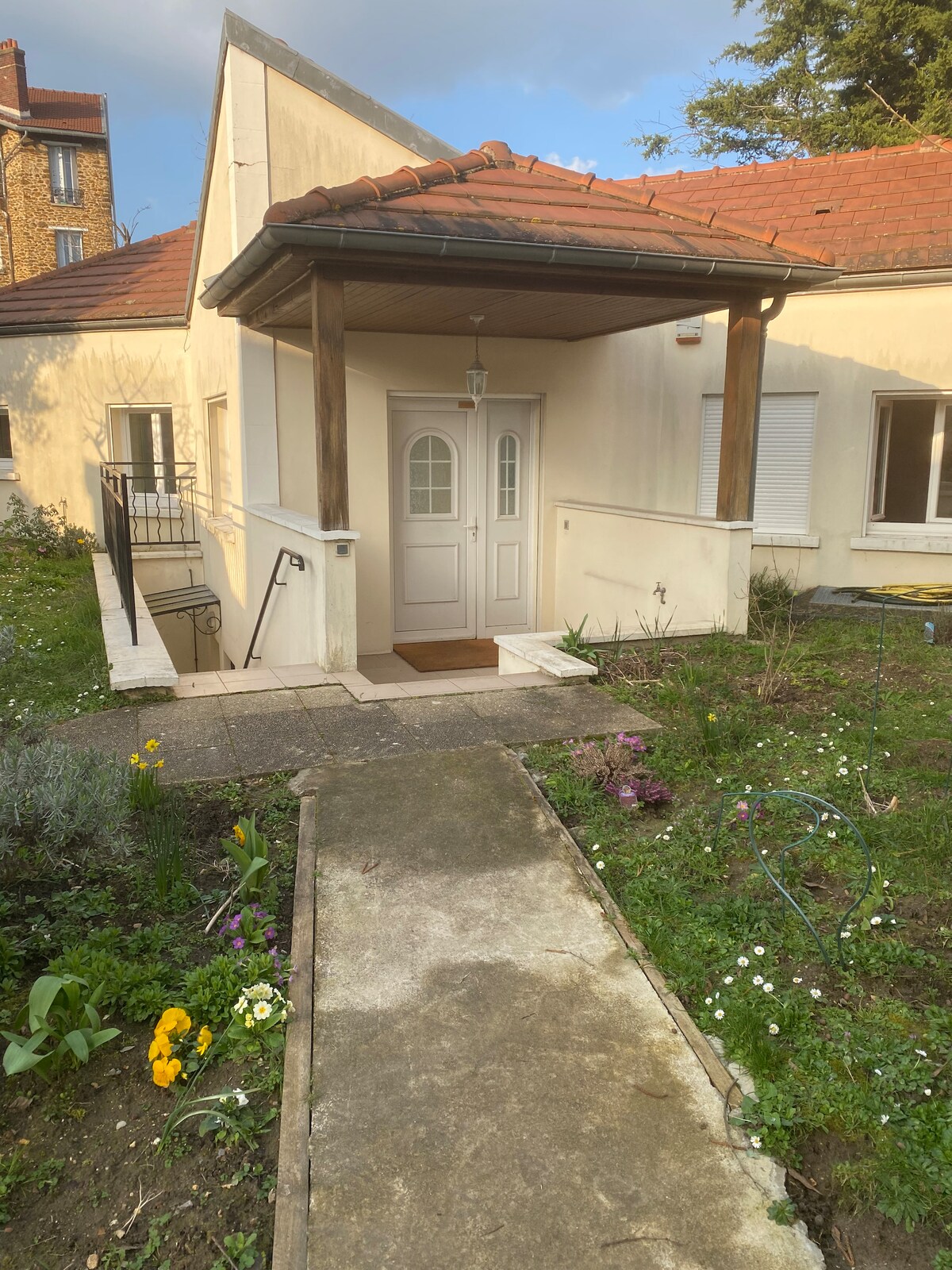 The entrance features a covered porch with a double door, framed by wooden posts. A stone pathway leads to the door, bordered by greenery and seasonal flowers, creating a welcoming approach to the home.
