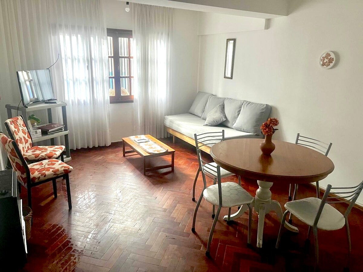 A living area is presented with natural light filtering through sheer curtains. A gray sofa is arranged next to a coffee table with decorative items. A round dining table is surrounded by metal and wooden chairs, while a television stands on a stand nearby.