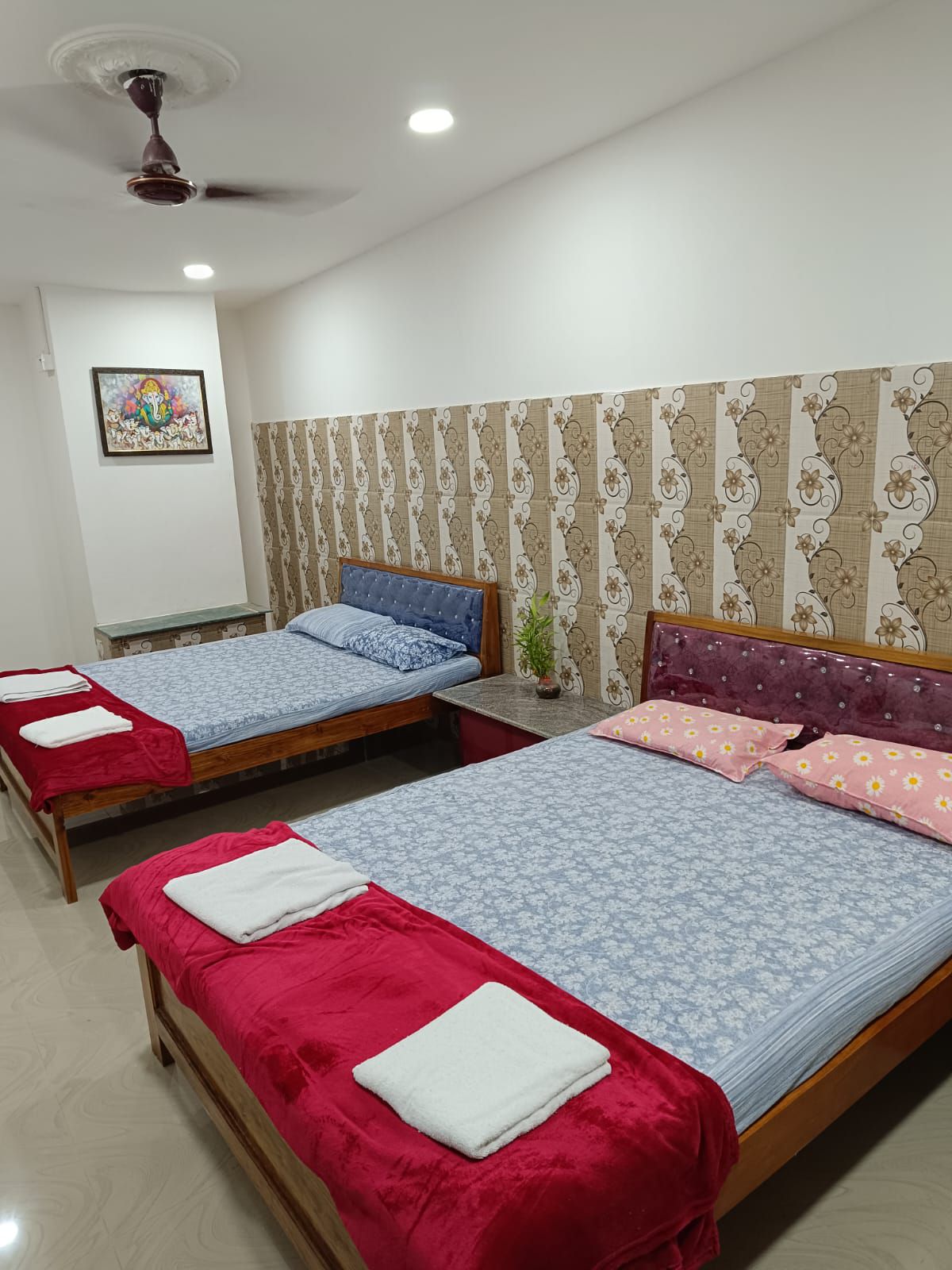 A well-lit room features two wooden beds, each with a blue floral bedding set and vibrant pink pillows. Red blankets are neatly placed at the foot of each bed. A small bedside table stands beside the beds, with a decorative piece visible on the wall.