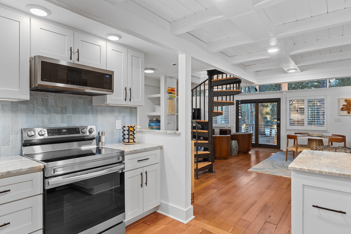 A modern kitchen is highlighted with white cabinetry and stainless-steel appliances. Light wood flooring extends into an open living area, featuring spiral stairs and large windows letting in natural light. A casual dining space is visible, adorned with a soft rug and contemporary furniture.