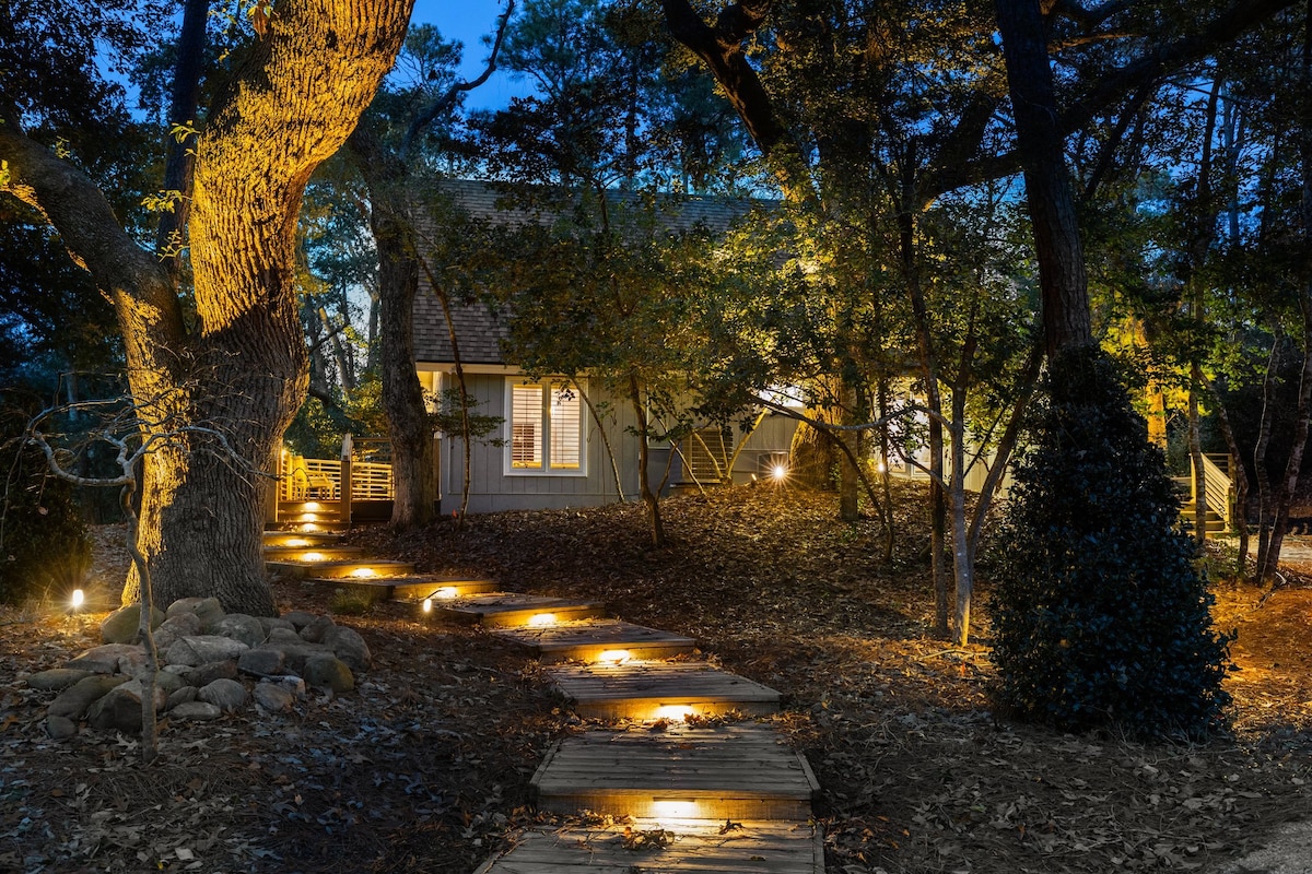 A winding wooden pathway is illuminated by soft lighting, leading through a serene garden area surrounded by lush trees. The welcoming exterior of the home is visible, with large windows revealing glimpses of the interior.