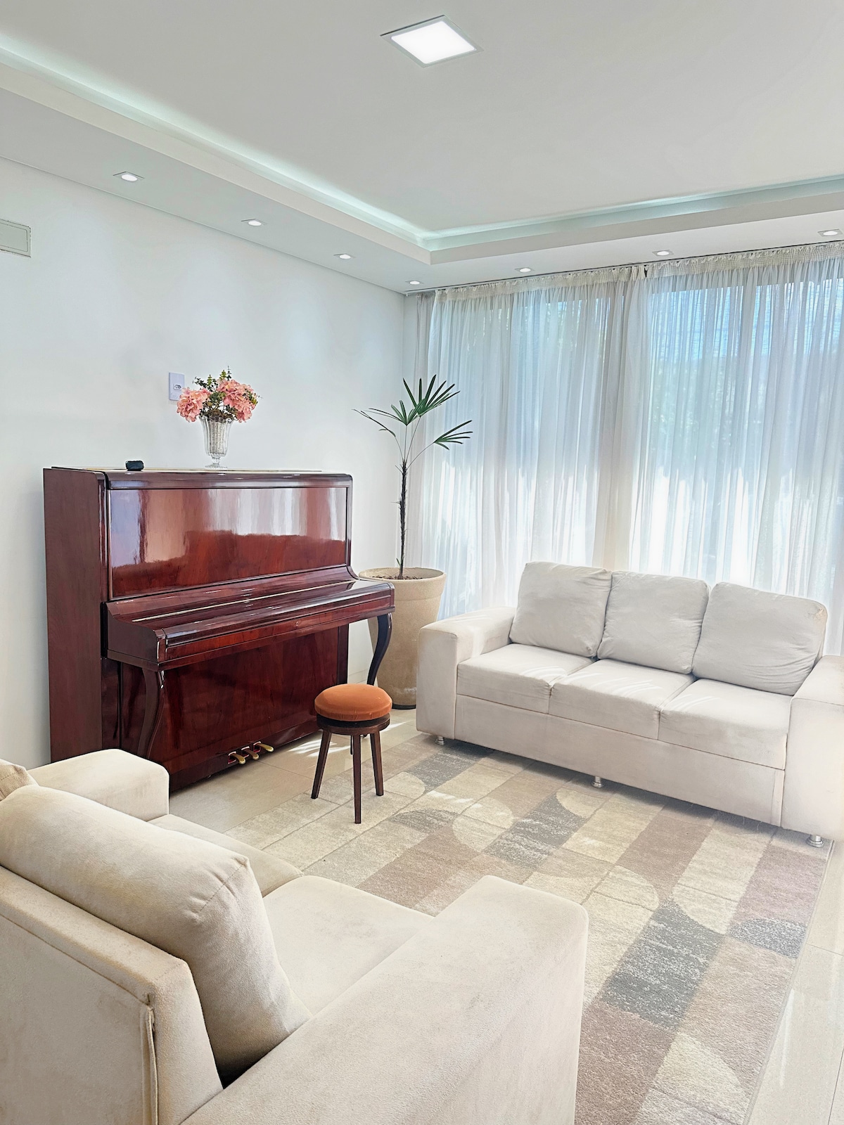 A living room features a polished wooden piano against a wall, complemented by a small decorative vase with flowers on top. Two light-colored sofas provide comfortable seating arrangements, while natural light streams in through large, sheer curtains, illuminating a patterned area rug.