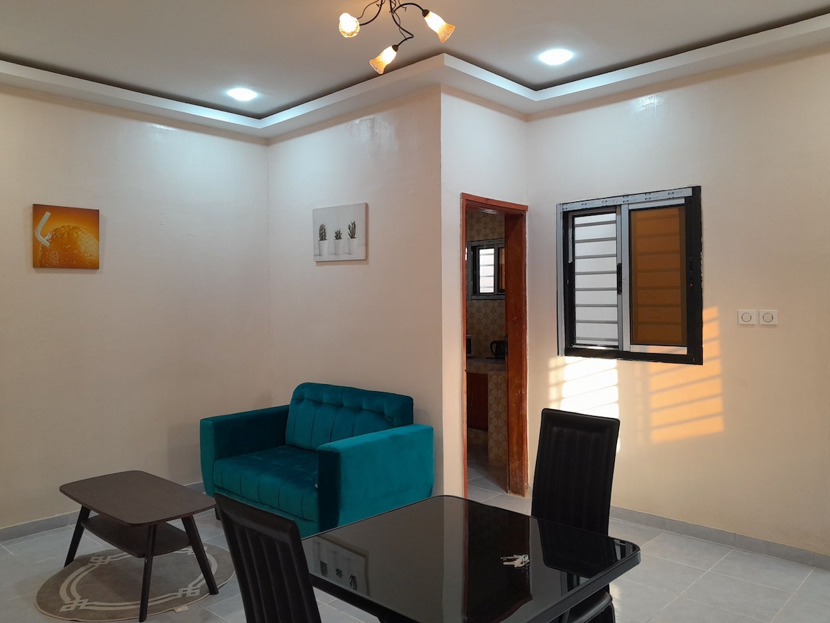 A contemporary living space features a plush blue sofa and a dark dining table with four chairs. Natural light filters through the window, illuminating the beige walls. A doorway leads to a separate area, with a small glimpse of additional interior visible.