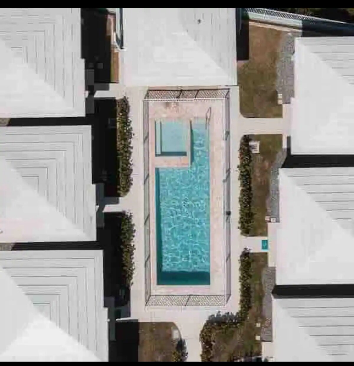 Aerial view of a shared rectangular pool nestled between two rows of villas. The pool features a shallow area, with surrounded sun loungers and landscaped greenery enhancing the setting. The villa roofs, designed in a symmetrical pattern, reflect a modern architectural style.