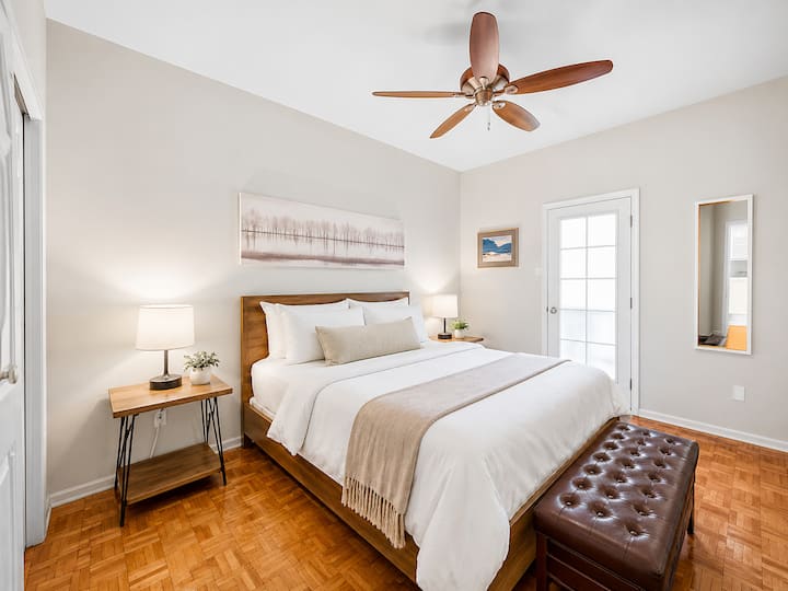 Une chambre lumineuse et confortable avec un grand lit, des lampes de chevet et un banc douillet au pied du lit. La chambre offre une ambiance calme et reposante avec un décor simple et un accès pratique par la porte privée avec des stores occultants.