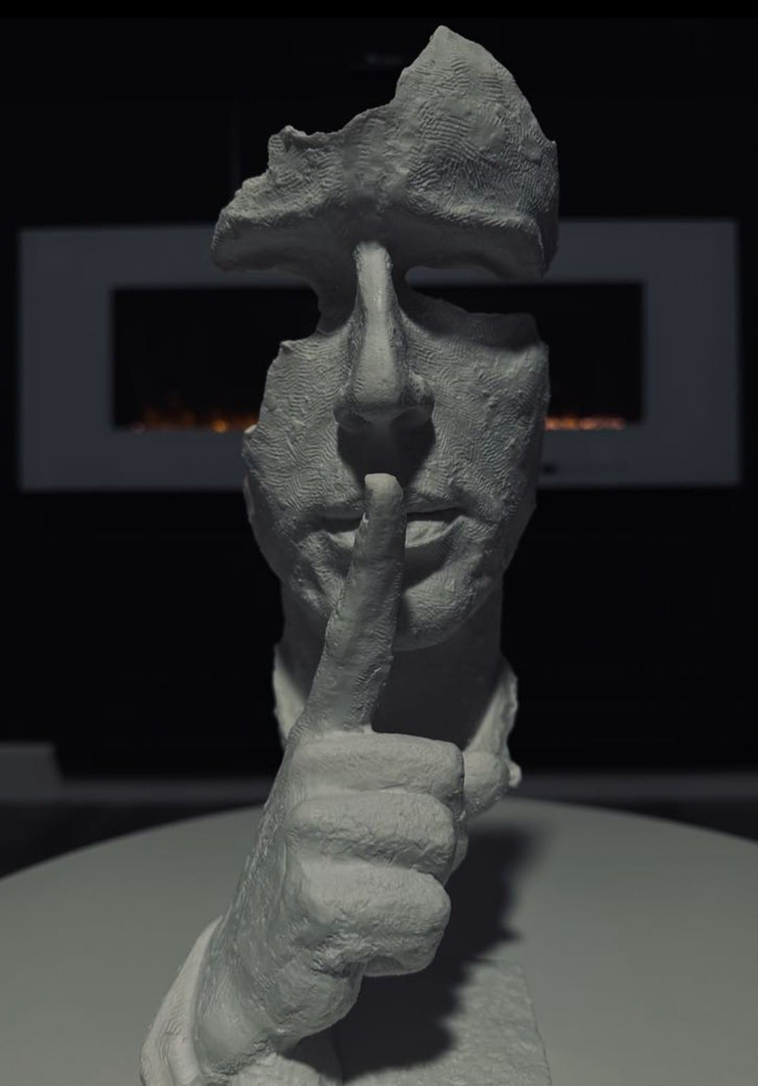 A white sculptural figure is depicted with a raised finger positioned to its lips, suggesting silence. The background features dark walls and what appears to be two modern fireplace units, providing a contrasting element to the light-toned sculpture.