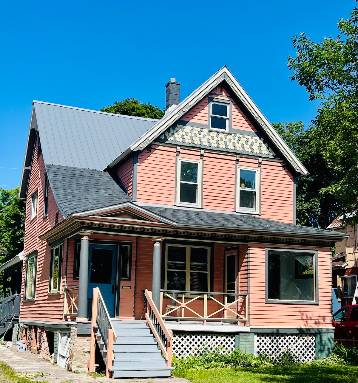 The exterior of a Victorian house features a pink facade with decorative trim and a signature gabled roof. Large windows allow natural light to fill the rooms, while a welcoming front porch is accessed by a set of steps. Lush greenery surrounds the property.
