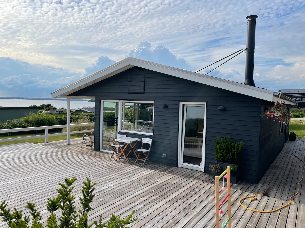 A modern summer house with a charcoal exterior is showcased, featuring large windows that invite natural light. The expansive wooden deck, equipped with outdoor furniture, offers a tranquil space. A chimney suggests a cozy wood-burning stove inside, while greenery surrounds the terrace.