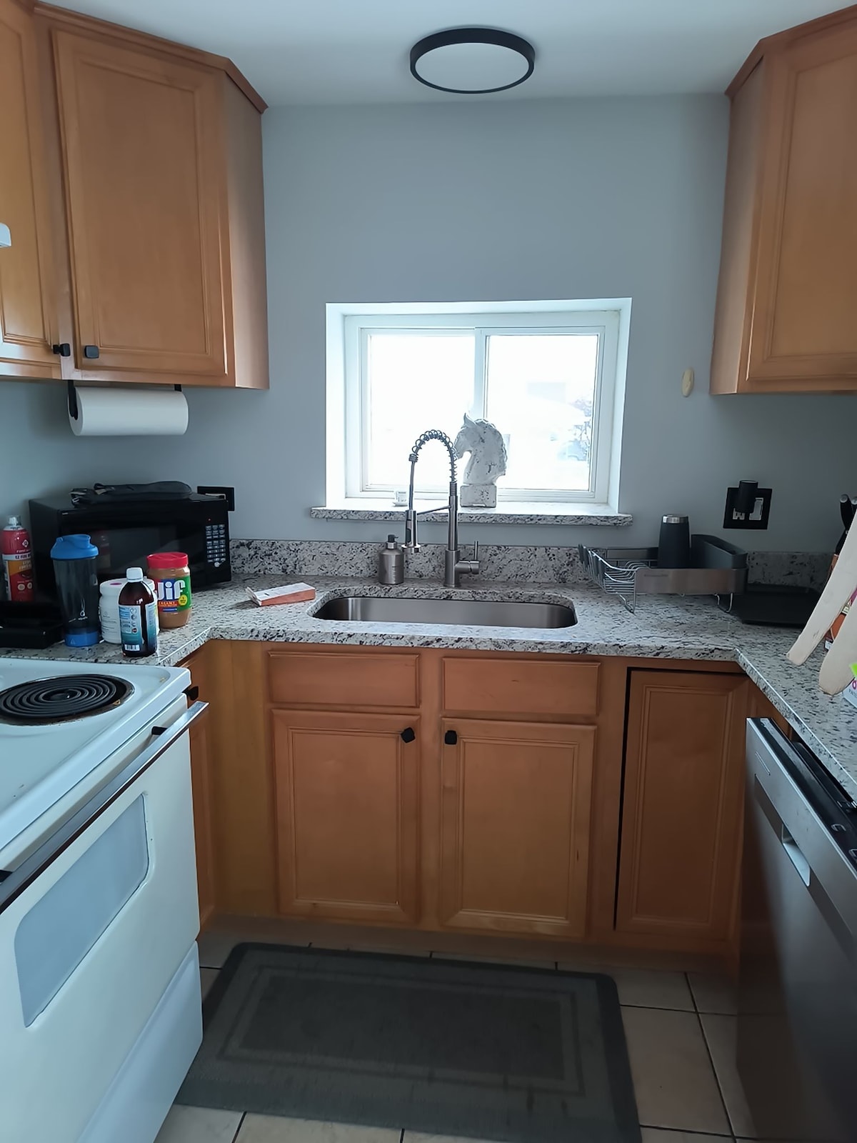 A compact kitchen features warm wooden cabinetry and granite countertops. A stainless steel sink is positioned beneath a window, allowing natural light to illuminate the space. Essential appliances, including a stove and microwave, are neatly integrated within the layout.