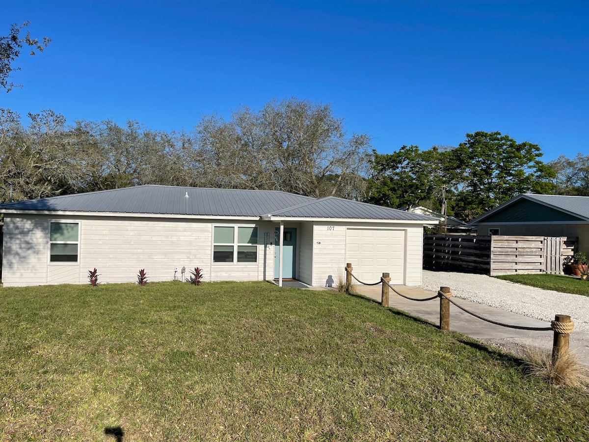 A single-story home is set in a spacious yard with well-maintained grass. The exterior features a light-colored facade and a carport. A circular gravel driveway is visible, surrounded by a rope boundary. Lush greenery forms a serene backdrop, enhancing the inviting ambiance.