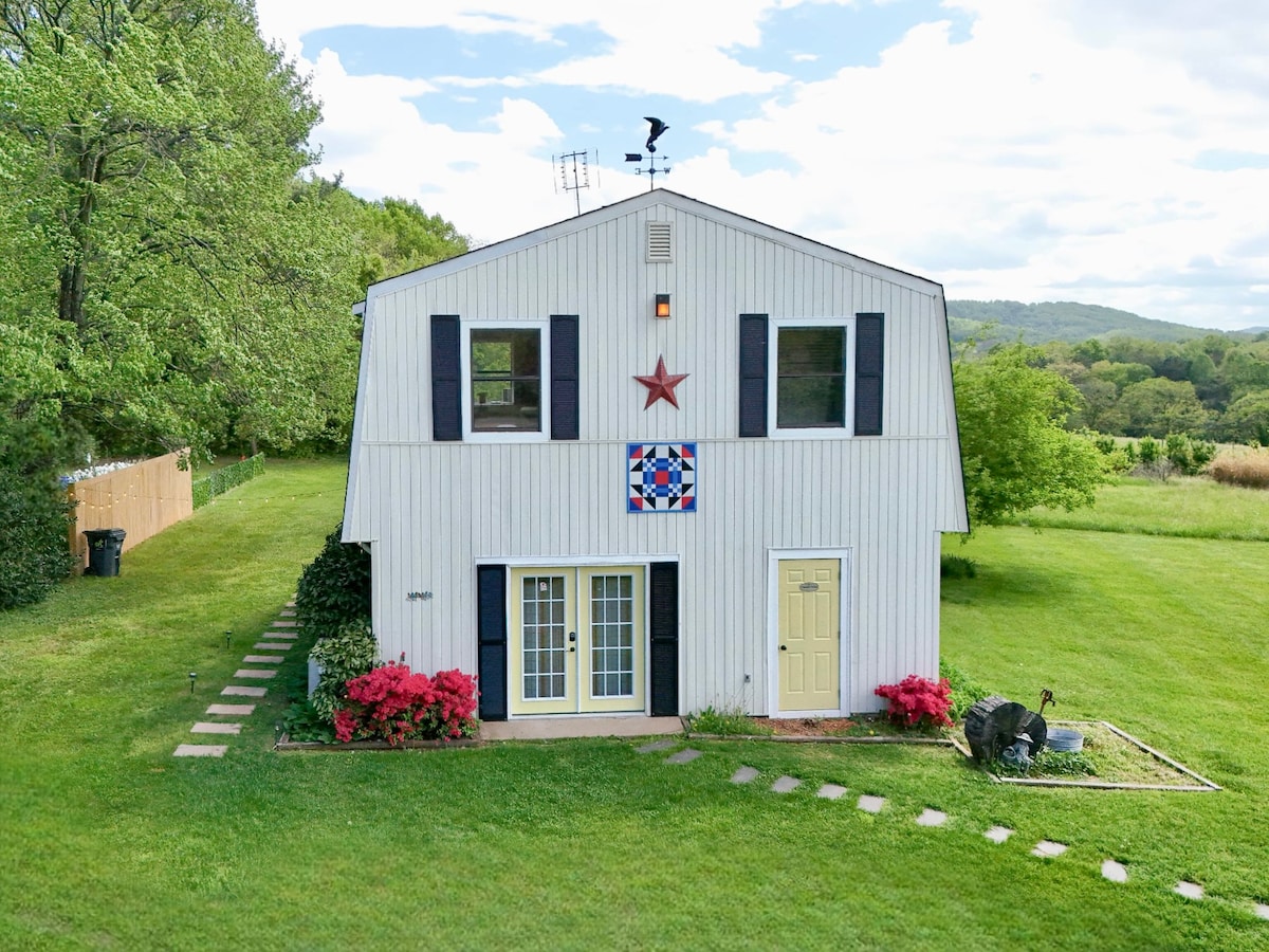 The exterior of a two-story barndominium is captured, featuring a white facade with blue shutters and a decorative red star. A lush green lawn surrounds the building, complemented by vibrant floral accents and stone pathways leading to the entrance.