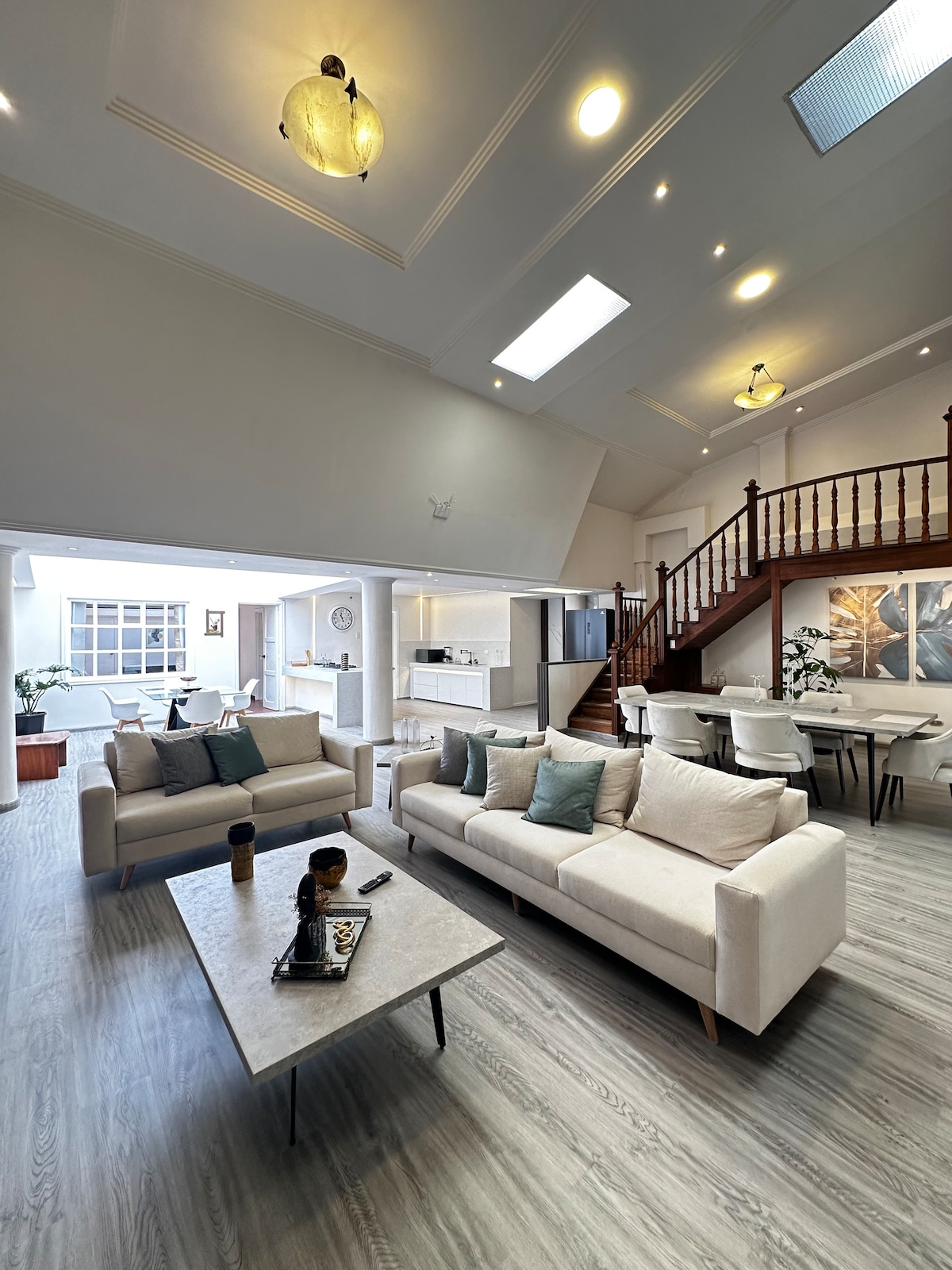 The living area features two light-colored sofas arranged around a central coffee table. Soft lighting illuminates the spacious room, which boasts high ceilings and large windows. A staircase with wooden railing leads to an upper level, while the dining area is positioned in the background.