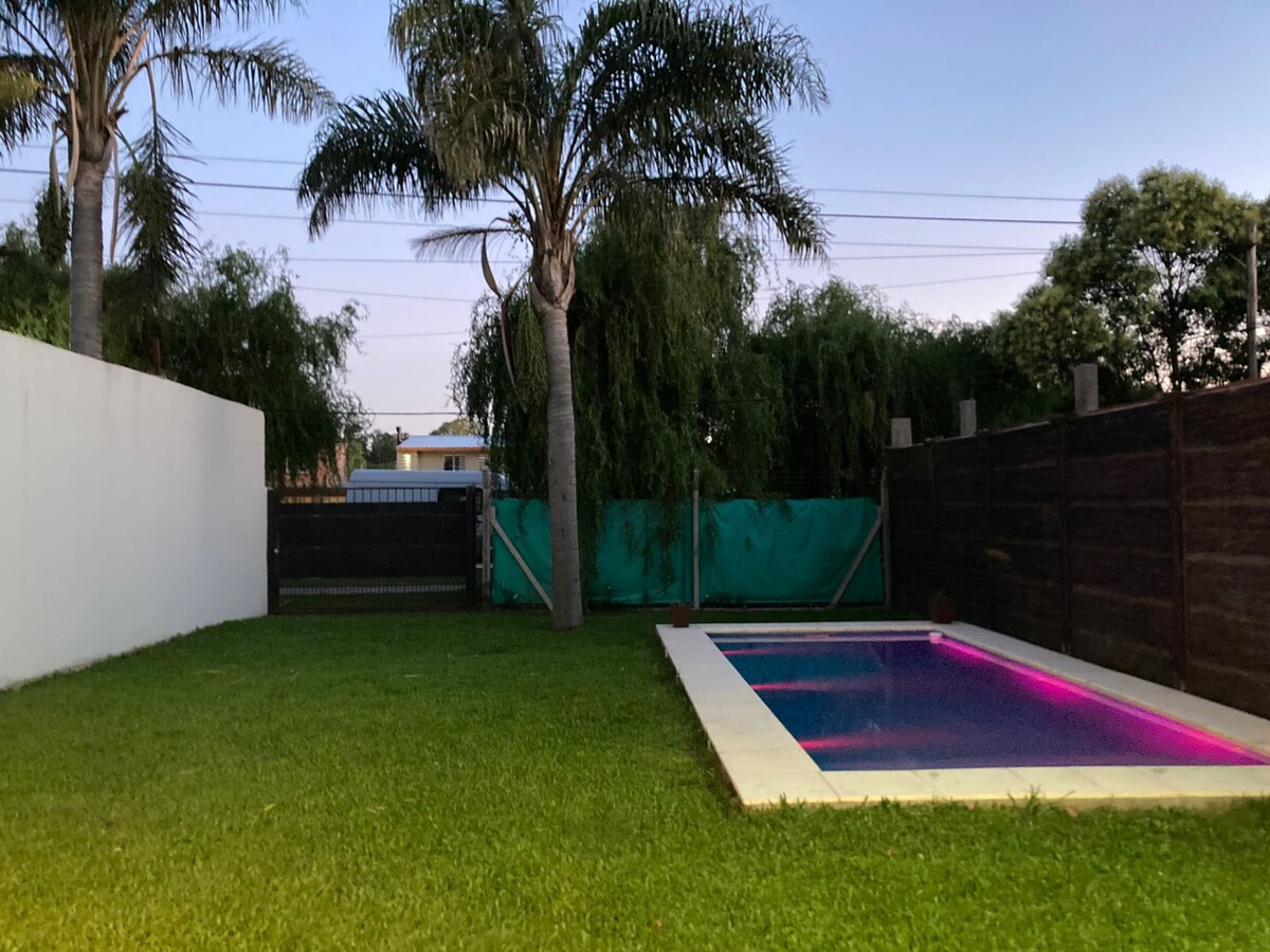 An outdoor area features a swimming pool with adjustable lighting, surrounded by lush green grass. Tall palm trees provide shade against a backdrop of a white wall and wooden fencing, enhancing the serene ambiance of the space.