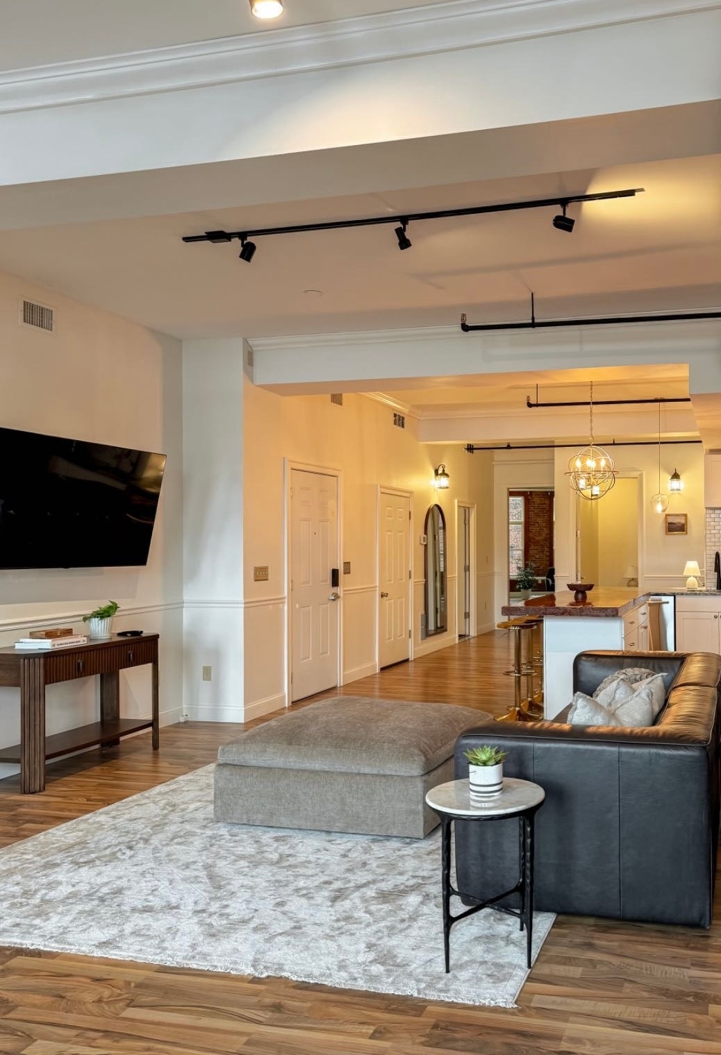 A spacious living area features a gray sectional sofa and a plush area rug. A mounted television is visible on the wall, complemented by a side table with a small potted plant. Warm lighting and wooden flooring enhance the inviting atmosphere.