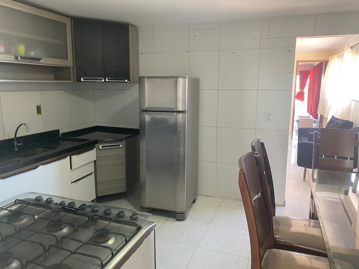 A modern kitchen is presented, featuring stainless steel appliances, including a refrigerator and a gas stove with an oven. The cabinets have a sleek design, and a dining area with brown chairs is visible in the background, illuminated by natural light.