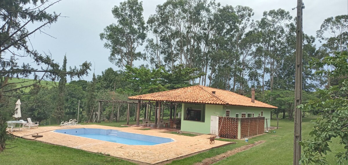 A spacious house is set amidst greenery, featuring a terracotta tiled roof. An outdoor pool lies in front, surrounded by a stone patio. Tall trees provide shade, enhancing the serene rural atmosphere. The property includes a covered area with seating options and is situated on a lush lawn.