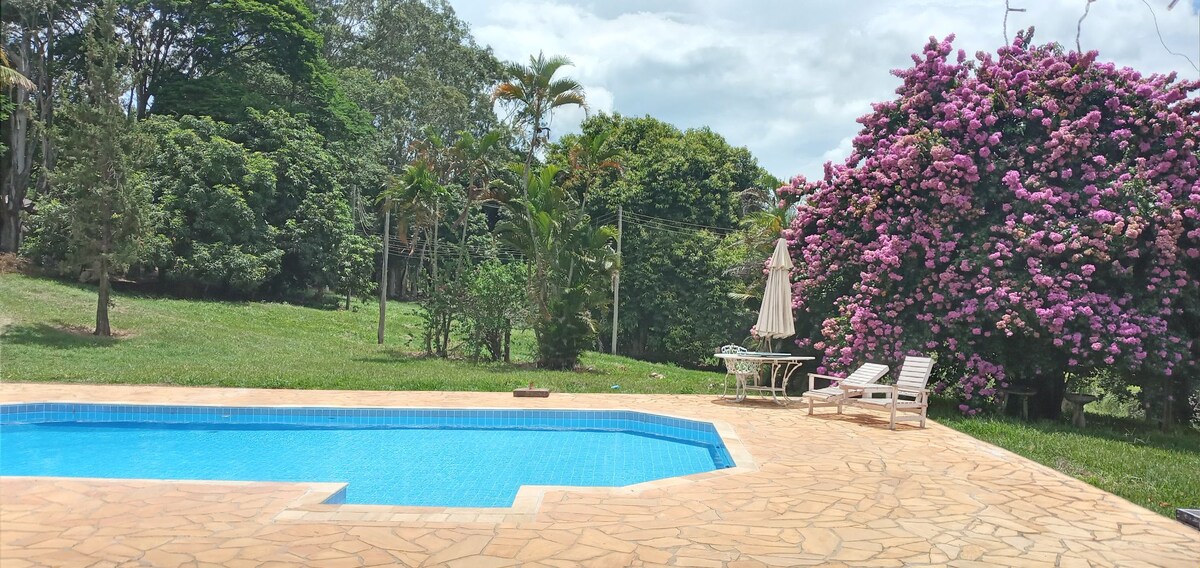 A serene pool area is bordered by a textured stone patio, featuring a refreshing blue pool. Lush greenery and vibrant flowering plants surround the space, while a sun umbrella shades a small table and chairs positioned nearby.