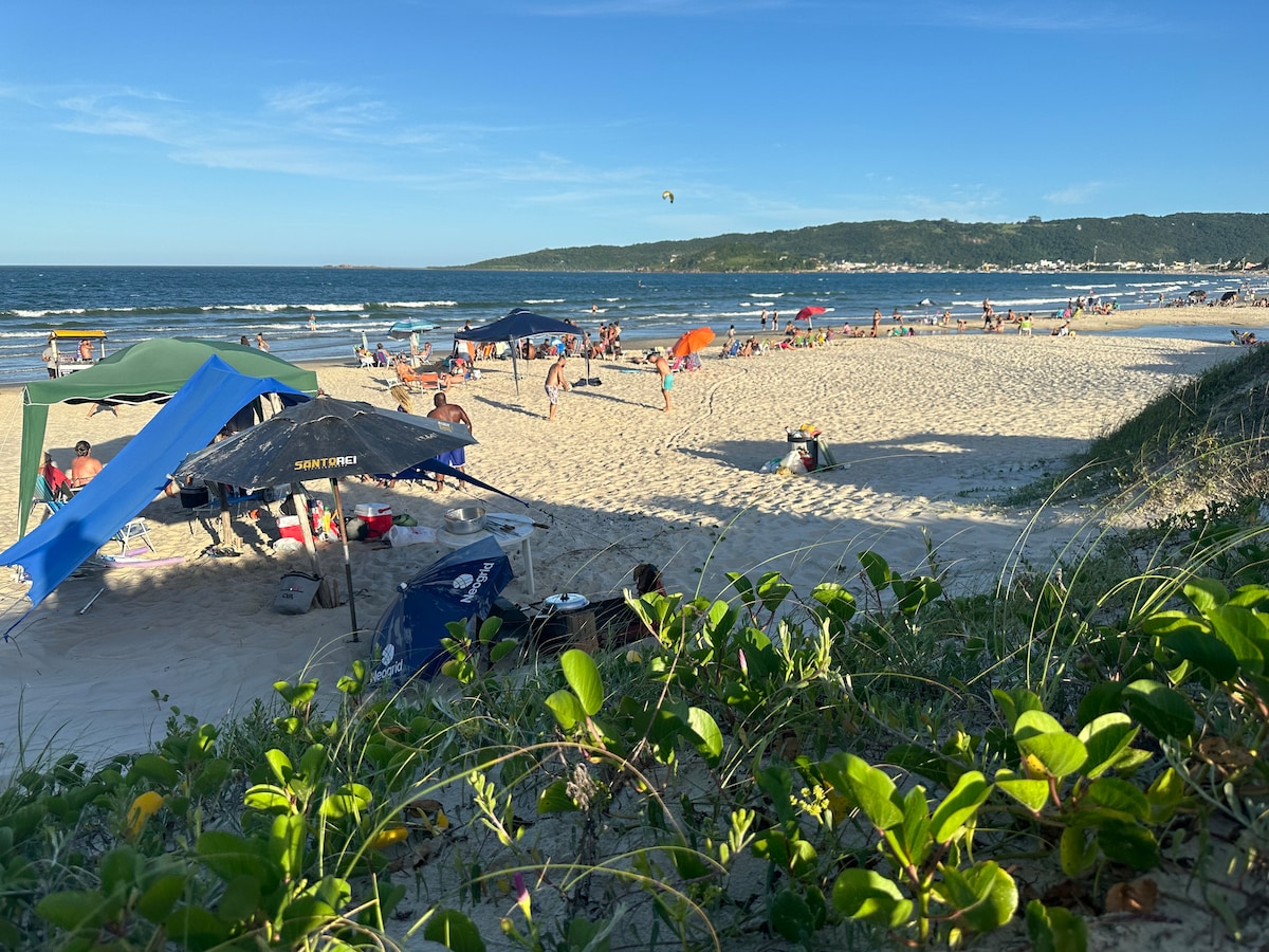 A sandy beach scene is captured, filled with people enjoying the sun. Colorful umbrellas provide shade for visitors, while gentle waves ripple in the background. Lush greenery frames the foreground, creating a natural border around the lively beach atmosphere.