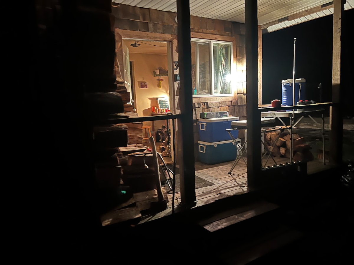 The exterior of a cabin is depicted at night, showcasing a well-lit porch area with a table and chairs. A cooler and water jug are visible, suggesting an outdoor dining setup. The warmth of light spills out from the cabin's interior, enhancing the welcoming atmosphere.