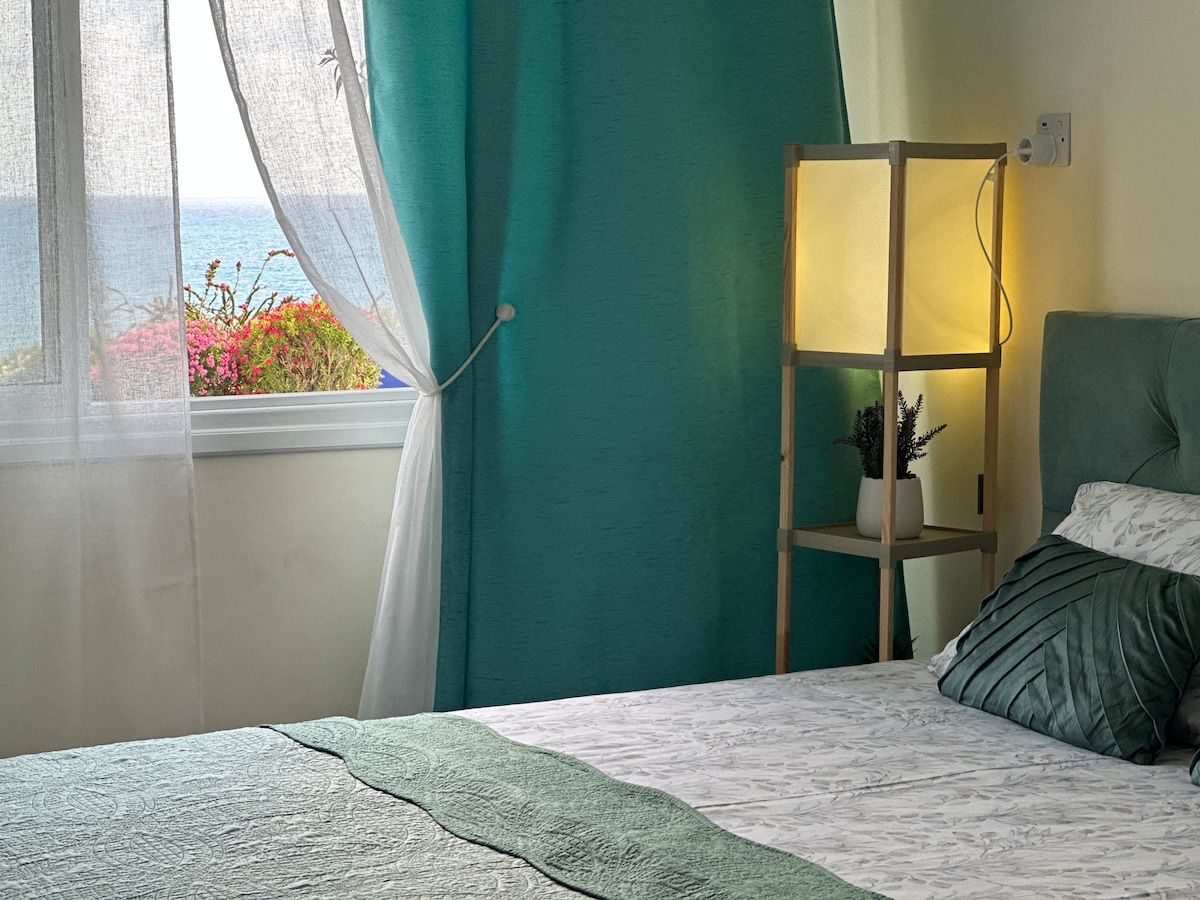 A cozy sleeping area is presented, featuring a spacious bed adorned with a textured green throw. A soft lamp stands beside the bed, casting a warm light. Sheer curtains filter natural light, revealing views of the sea and colorful plants outside the window.