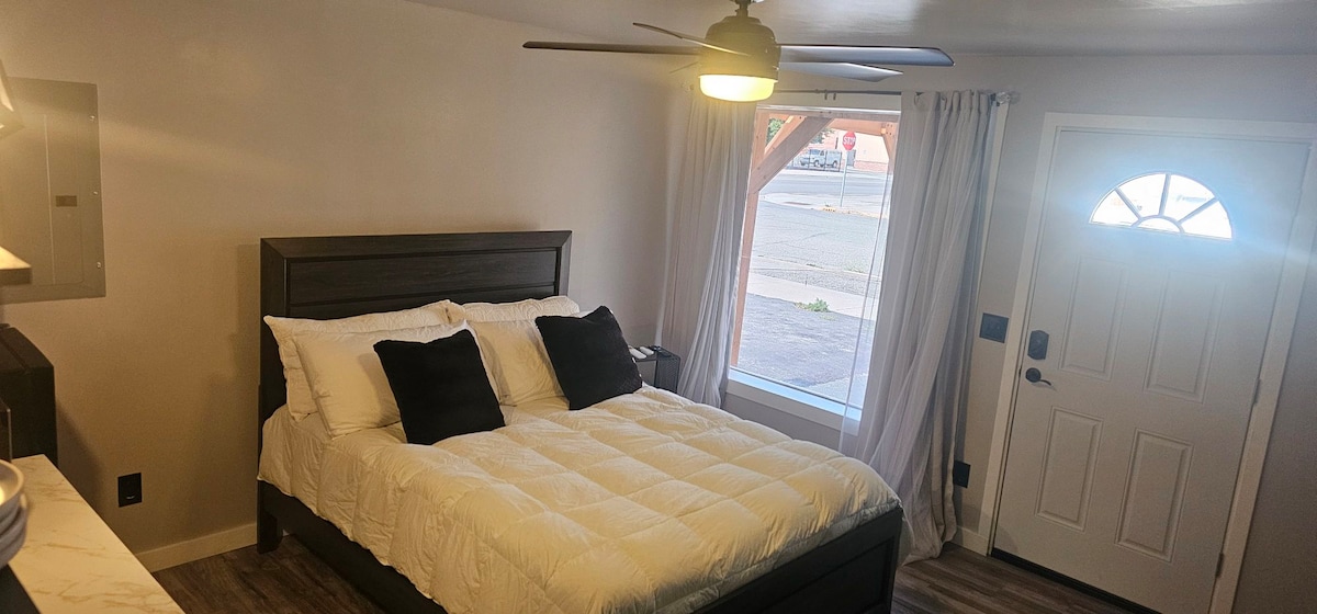 A cozy bedroom features a queen bed with white bedding and black accent pillows. Natural light enters through a nearby window, and a ceiling fan is installed for comfort. The entry door adds accessibility with a welcoming design.