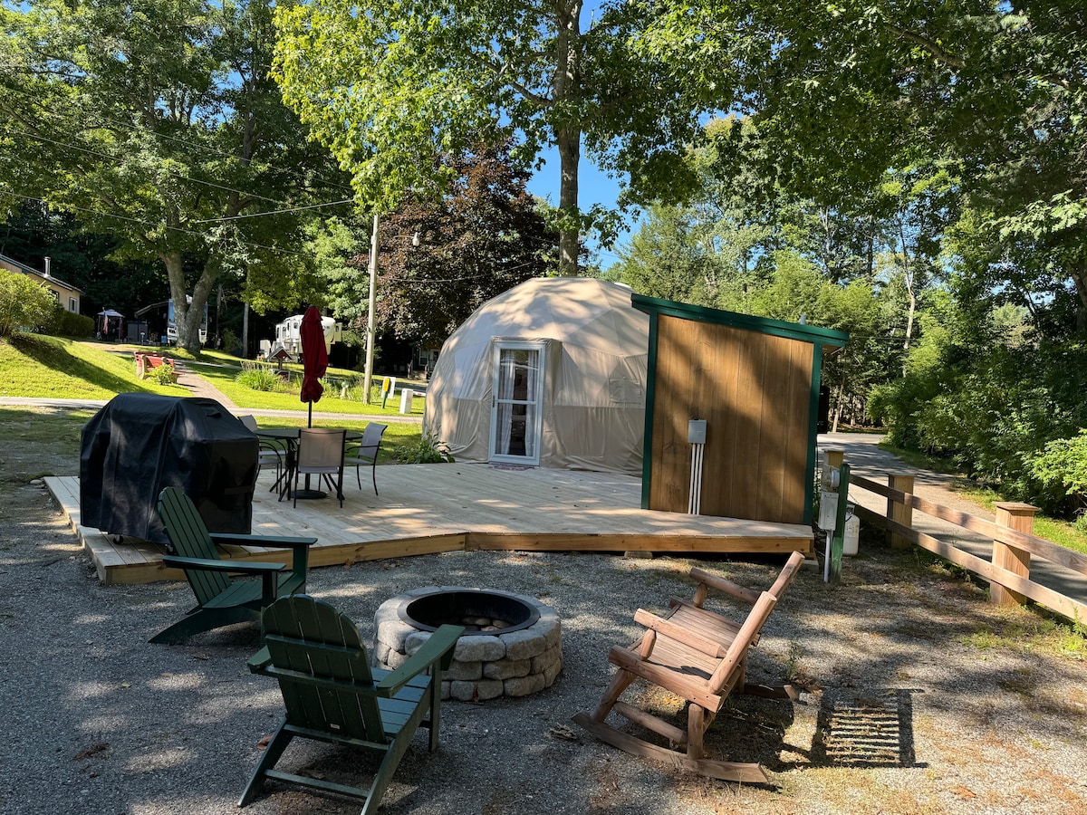 A Geo Dome is shown with a spacious deck featuring a table and chairs. A barbecue grill is positioned nearby, along with a fire pit surrounded by seating. Tall trees provide shade, and the campground area is visible in the background.