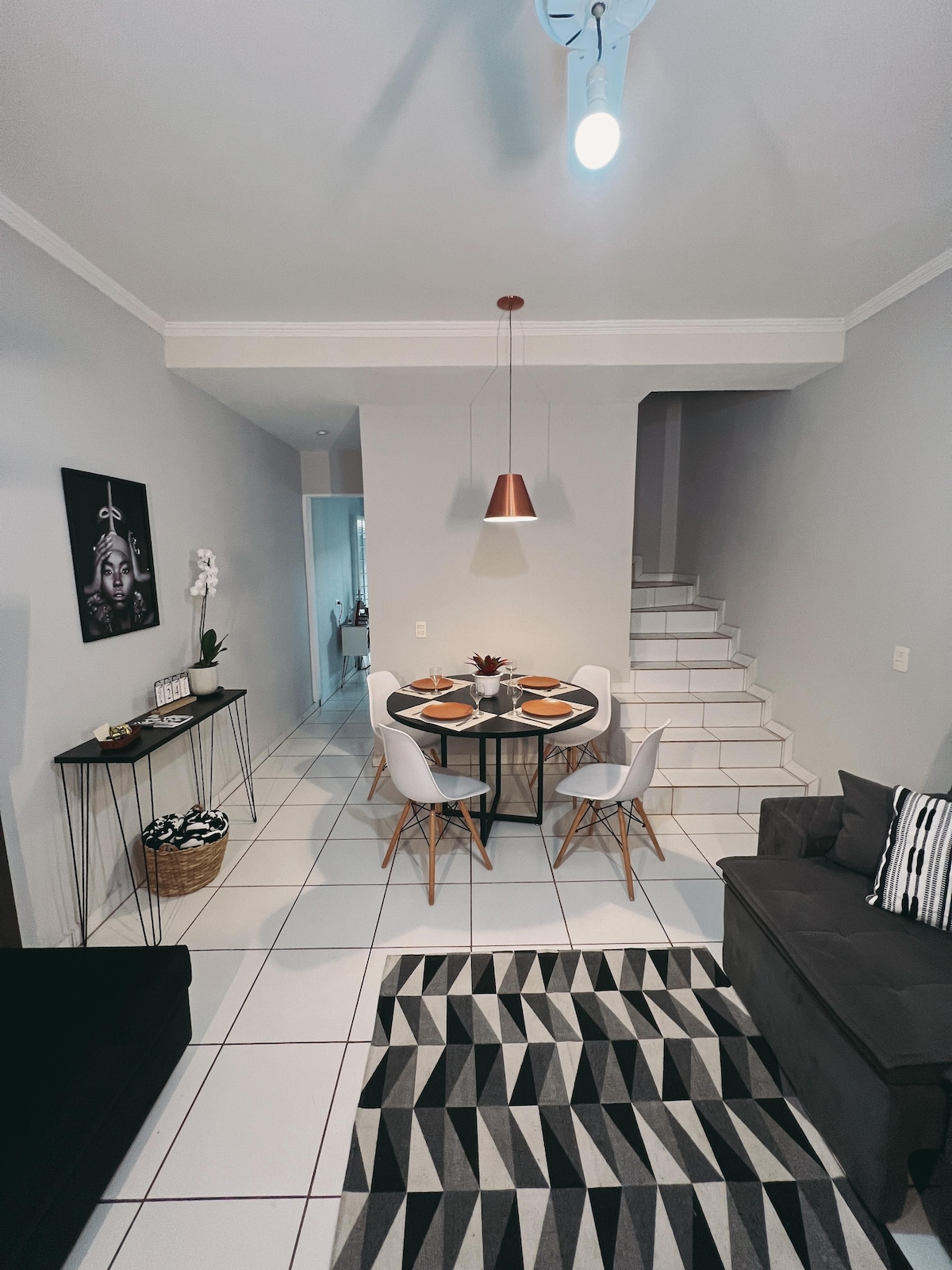 A dining area is positioned between a staircase and a cozy living space. A round table is set for two, surrounded by modern chairs, while a geometric rug adds texture to the tiled floor. Soft lighting is provided by a pendant lamp above the table.