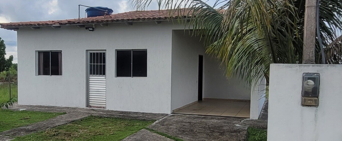 The exterior of the house features a simple white façade with two large windows and a door, framed by well-maintained grass and palm trees. A concrete pathway leads to the entrance, while a water tank sits on the roof, blending functionality with the structure's design.
