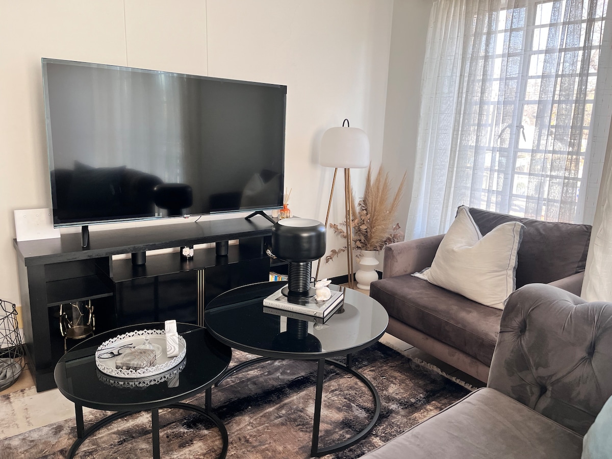 A modern living area is presented with a large flat-screen television mounted on a sleek black unit. A round coffee table sits in the center, surrounded by a plush gray sofa and a contemporary armchair. Natural light filters through sheer curtains, illuminating decorative accents.