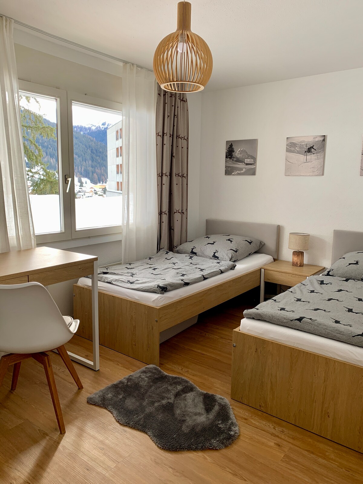A bright sleeping area features two individual beds with patterned bedding. A small desk and chair are positioned nearby, while a soft rug adds warmth to the wooden floor. Light enters through a large window, offering views of the surrounding mountains.