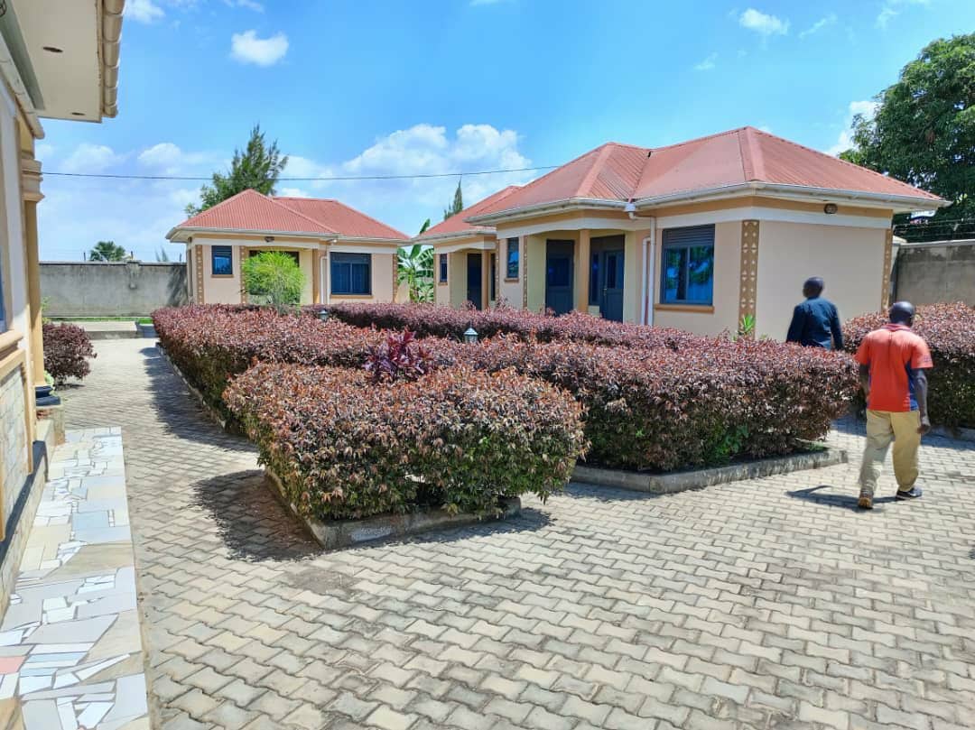 A serene outdoor space is presented, featuring two charming cottages with red roofs and blue window frames. Lush green bushes line the pathway, creating a natural border between the cottages, while clear skies enhance the tranquil atmosphere.