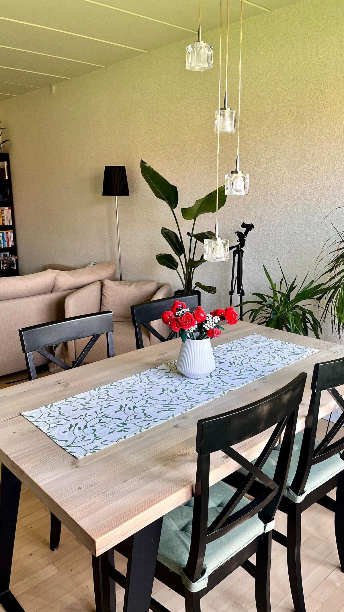 A dining table is centered in the image, adorned with a floral table runner and a vase of red roses. Surrounding chairs provide seating for guests. Soft light illuminates the room, with greenery from plants enhancing the inviting atmosphere. A floor lamp stands in the background.