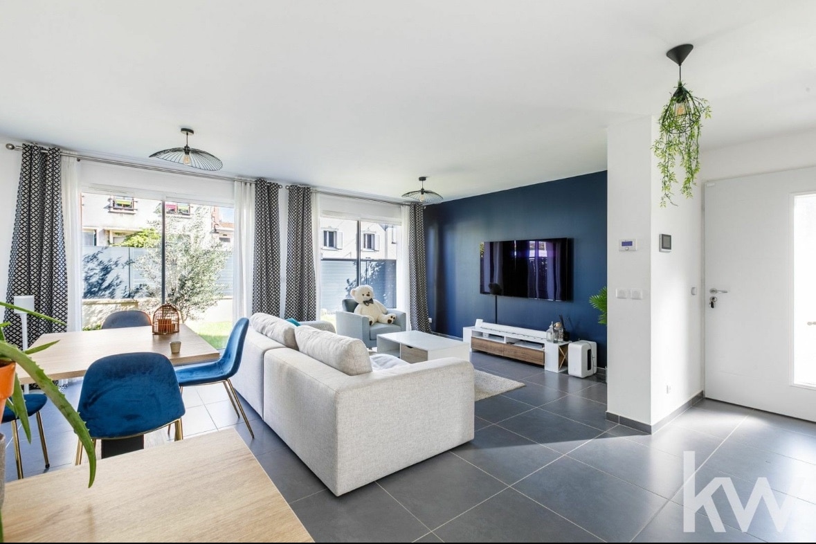 A bright living space features a spacious light-colored sofa and a sleek entertainment unit positioned against a dark blue wall. Large windows invite natural light, while a dining area, furnished with a wooden table and blue chairs, is visible in the background.