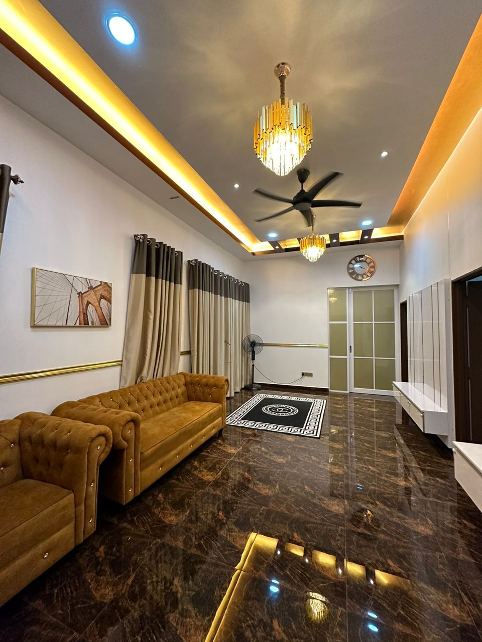 A spacious living area features elegant lighting, highlighting dark marble flooring and a plush, tan-colored sofa. Tall curtains frame the windows, and a decorative clock is mounted on the wall. A fan hangs from the ceiling, contributing to a comfortable ambiance.