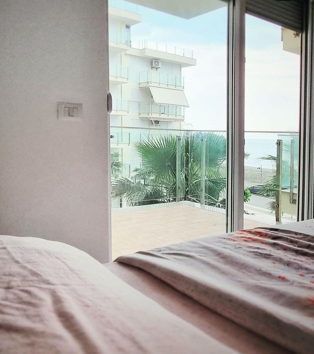 A view from a cozy sleeping area showcases a balcony with glass railings and tropical plants. The bright ambient light enters from the large sliding doors, revealing the serene beach scenery beyond.