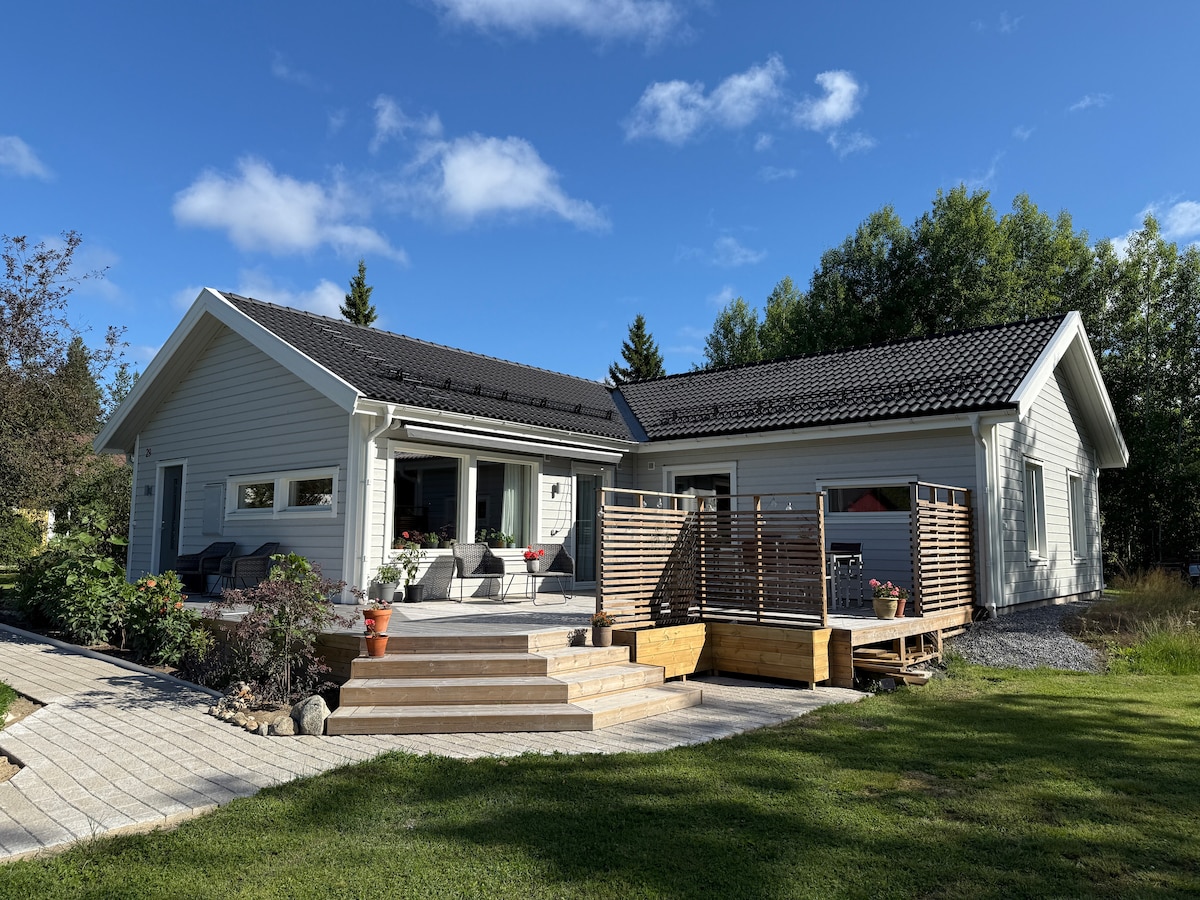 A modern house is depicted, featuring a spacious patio with a wooden railing and steps leading down to a lush lawn. The home's exterior is accented by large windows and a gray roof against a backdrop of greenery and blue skies.
